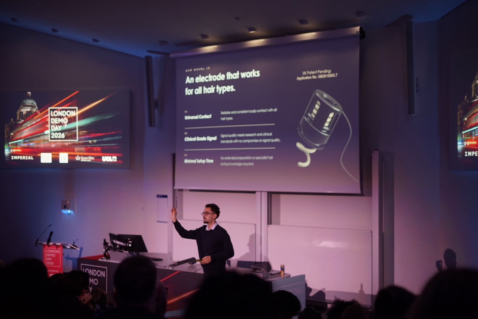 Merlin Angel Kelly presenting Syn-R1 on stage at London Demo Day 2026, in front of a seated audience, with a slide reading "An electrode that works for all hair types"